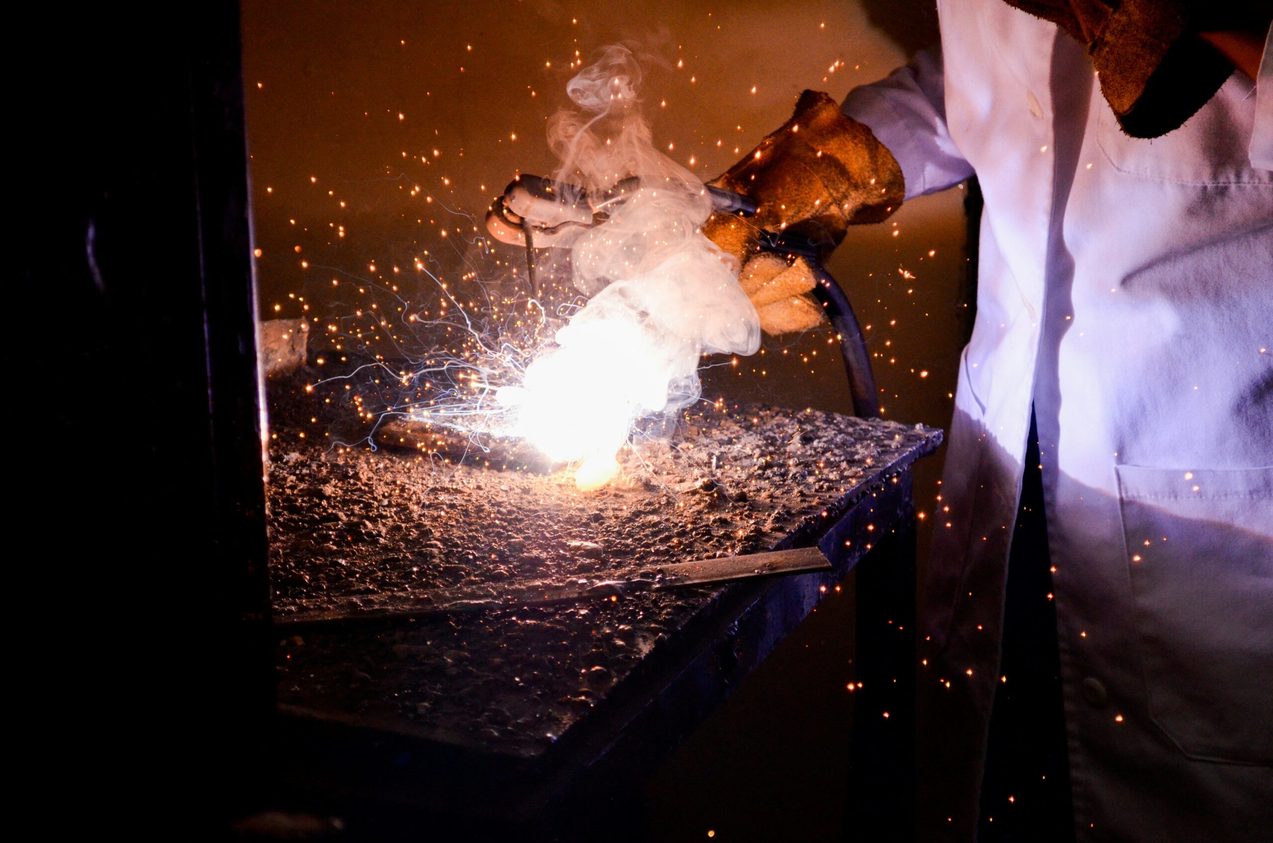Close-up of a person welding metal with bright sparks and smoke, wearing protective gloves and a white coat.

